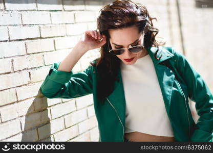 Young brunette woman with aviator sunglasses. Girl, model of fashion, wearing green modern jacket, standing in urban background.