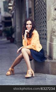 Young brunette woman with beautiful legs, model of fashion, wearing orange modern jacket and blue skirt, sitting in urban background.