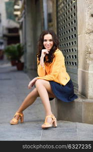 Young brunette woman with beautiful legs, model of fashion, wearing orange modern jacket and blue skirt, sitting in urban background.