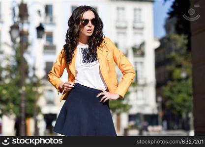 Young brunette woman with sunglasses. Girl, model of fashion, wearing orange modern jacket and blue skirt, standing in urban background.