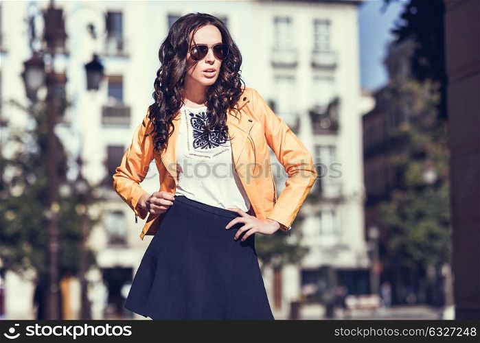 Young brunette woman with sunglasses. Girl, model of fashion, wearing orange modern jacket and blue skirt, standing in urban background.