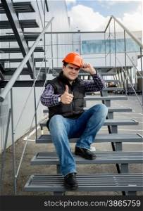 Young builder in orange helmet sitting on metal staircase and showing thumb up