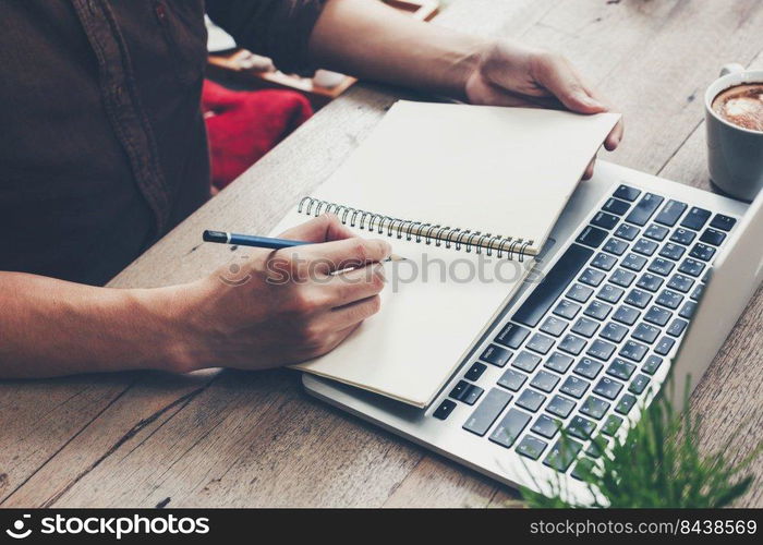 Young business man hand writing notebook and using laptop on wood table.