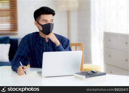 Young business man in face mask planning and working from home on laptop computer on desk for protection for outbreak of covid-19, male quarantine stay home using notebook on desk, new normal.