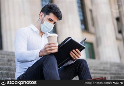 Young business man wearing a face mask and reading files while sitting on stairs outdoors. Business concept. New normal lifestyle concept.