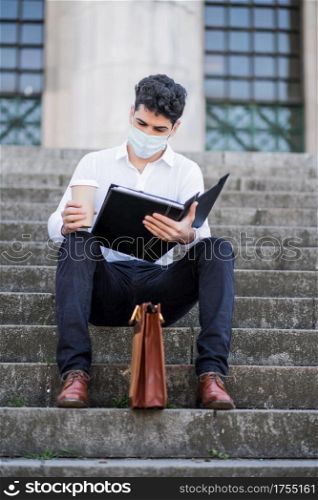 Young business man wearing a face mask and reading files while sitting on stairs outdoors. Business concept. New normal lifestyle concept.