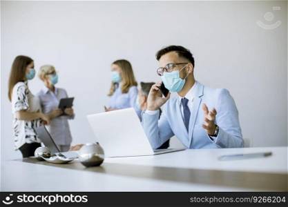 Young business man with medical protective mask works on a laptop and using mobile phone in the office