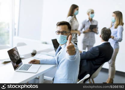 Young business man with medical protective mask works on a laptop in the office and showing positive thumb gesture