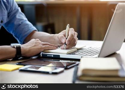 Young business man working in bright office, using laptop, writing notes.