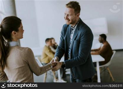 Young business partners making handshake in an office while their team working in the background
