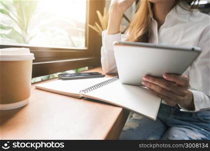 Young business woman in white dress sitting at table in cafe and writing in notebook. Asian woman using tablet and cup of coffee. Freelancer working in coffee shop. Student learning online.