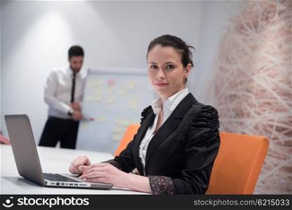 young business woman on meeting usineg laptop computer, blured group of people in background at modern bright startup office interior taking notes on white flip board and brainstorming about plans and ideas
