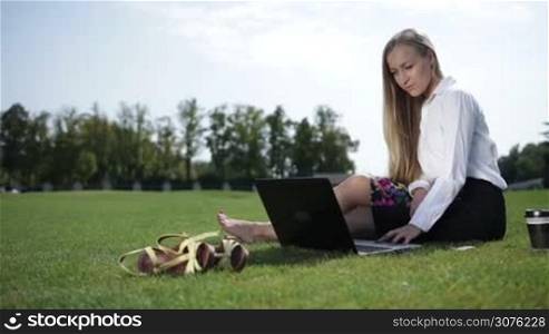 Young business woman sitting on grass barefoot in the park and working on laptop outdoor