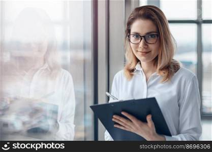 Young business woman standing at the window of a modern office and holding a folder with documents.