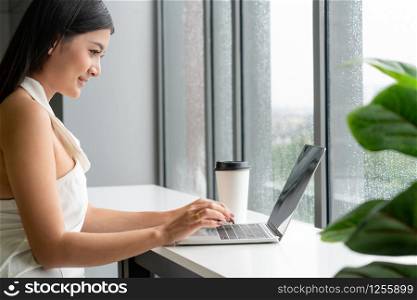 Young business woman using laptop computer while sitting at cafe table next to office windows.