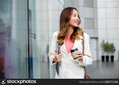 Young business woman wearing white suit jacket and smile while go to work outdoor mirror building background. Confident Businesswoman with cup of coffee holding Laptop walking outside office building