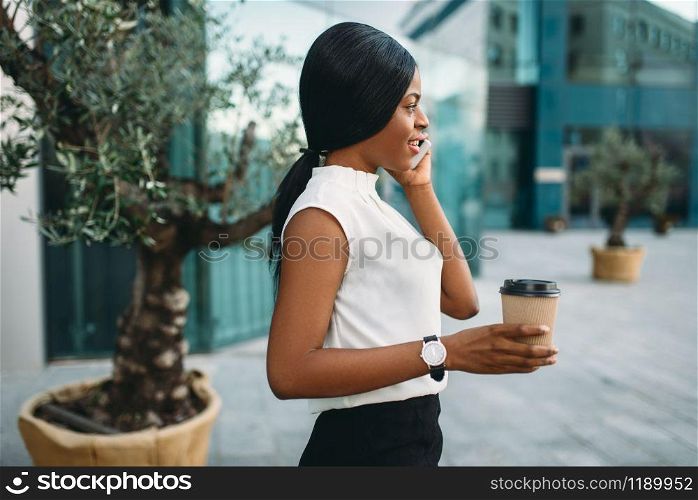 Young business woman with cardboard coffee cup and mobile phone against office building on background. Smiling black businesswoman in skirt and white blouse. Business woman with coffee cup and mobile phone