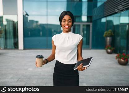Young business woman with cardboard coffee cup and notepad against office building on background. Smiling black businesswoman in skirt and white blouse