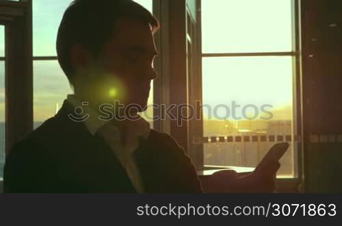 Young businessman at the airport. He using smart phone to type text message. Bright evening sunshine in the window