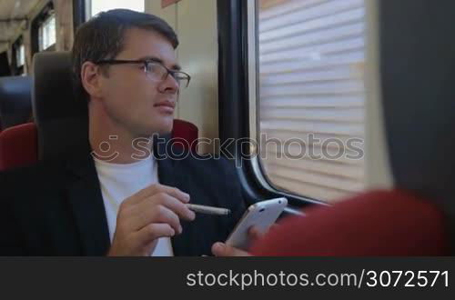 Young businessman browsing the internet on smart phone in train and enjoying view from the window with thoughtful look