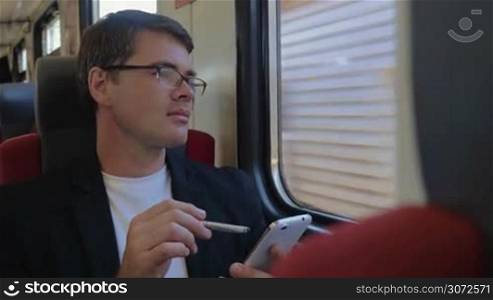 Young businessman browsing the internet on smart phone in train and enjoying view from the window with thoughtful look
