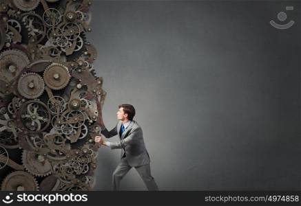 Young businessman carrying huge gears mechanism on back. Making mechanism work