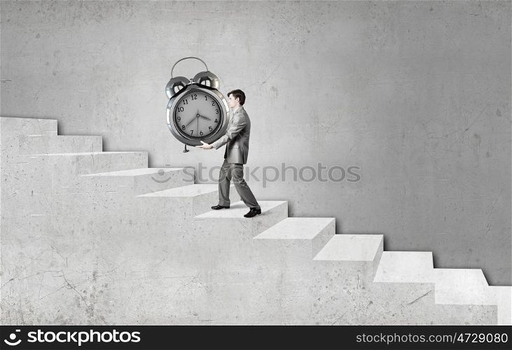 Young businessman carrying with effort huge alarm clock. Time concept
