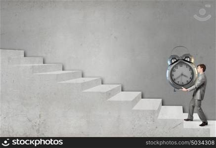 Young businessman carrying with effort huge alarm clock. Time concept