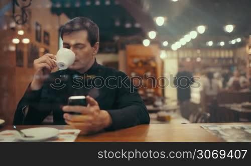 Young businessman drinking coffee and typing sms in a cafe. View through the glass with weak city reflection