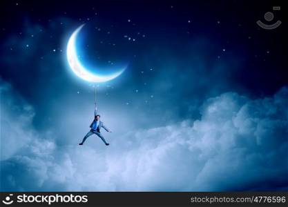Young businessman hanging on rope at night. Man hang on moon