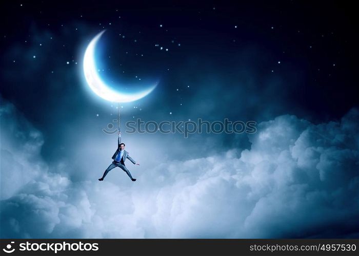 Young businessman hanging on rope at night. Man hang on moon