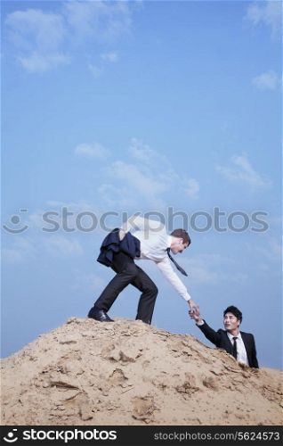Young businessman helping another businessman get to the top of the hill in the desert