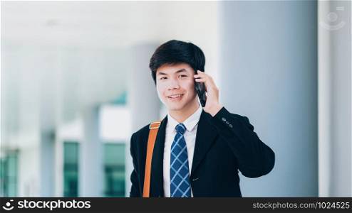 Young businessman in black suit speaking over mobile or smart phone with business partners and smile looking at the camera. Business outdoors.