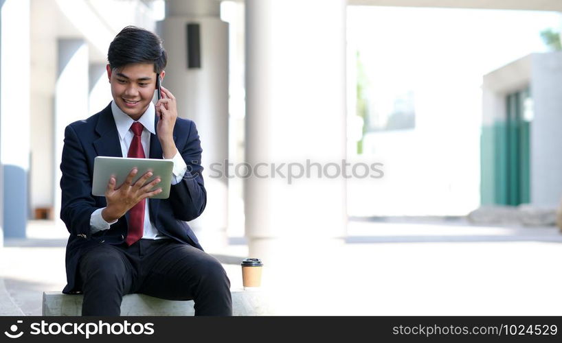 Young businessman in black suit speaking over mobile or smart phone with business partners. Business outdoors.