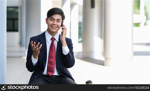Young businessman in black suit speaking over mobile or smart phone with business partners. Business outdoors.