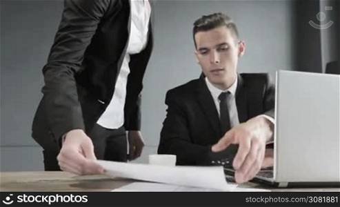 Young businessman in black suit using laptop in office, his collegue giving financial data diagram