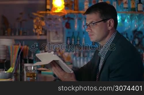 Young businessman in glasses and suit using tablet computer while sitting at the bar counter and having alcoholic drink