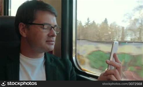 Young businessman in glasses having a video chat on smart phone while traveling by train