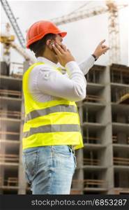 Young businessman in hardhat and safety vest talking by phone at construction site