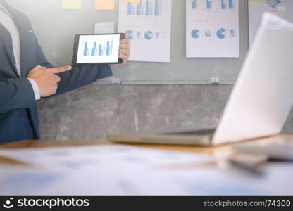 Young businessman in the suit explaining data chart on digital tablet and documents on the wall in a office.