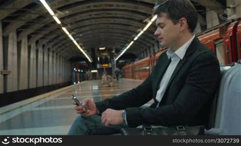 Young businessman is waiting for the tube train. He&acute;s passing the time with a smartphone.