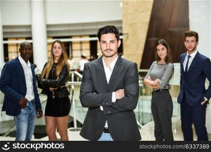 Young businessman leader looking at camera with arms crossed in office building. Group of muti-ethnic people in the background