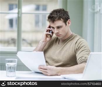 Young businessman looking at documents while using mobile phone in office