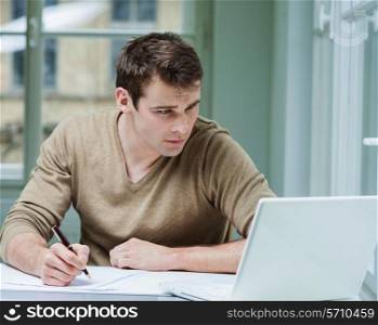 Young businessman looking at laptop while writing on documents in office