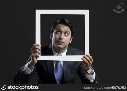 Young businessman looking up while holding picture frame over black background