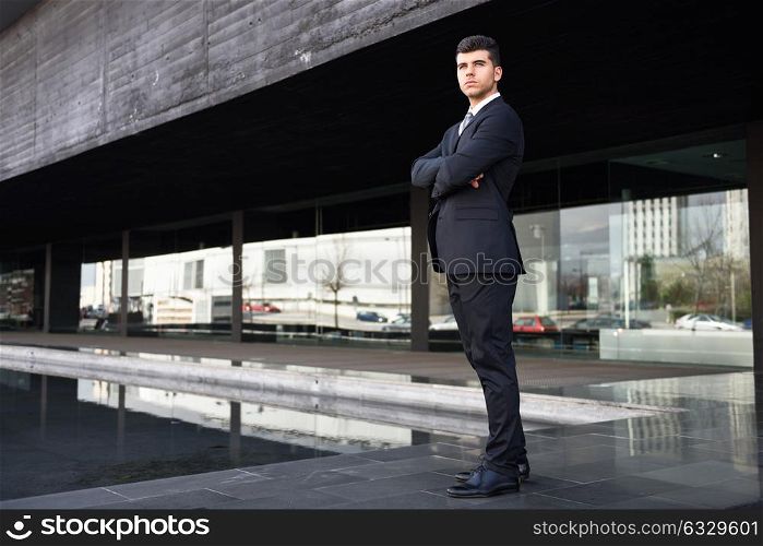 Young businessman near a modern office building wearing black suit and tie. Man with blue eyes
