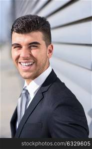 Young businessman near a modern office building wearing black suit and tie. Man with blue eyes smiling.