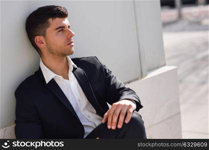 Young businessman near a modern office building wearing black suit and white shirt sitting on the floor. Man with blue eyes in urban background