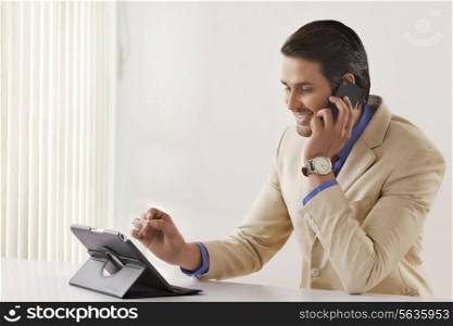 Young businessman on call while using digital tablet at office desk