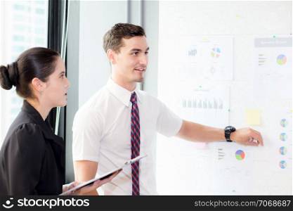 young businessman pointing towards graph and businesswoman holding clipboard with present profit while giving presentation in office, teamwork concept.